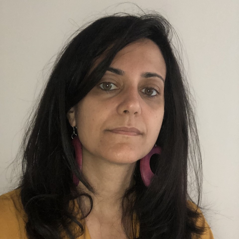 Estella Carpi. Woman with dark hair and striking magenta hoop earrings looking at the camera. She wears a mustard-yellow top.