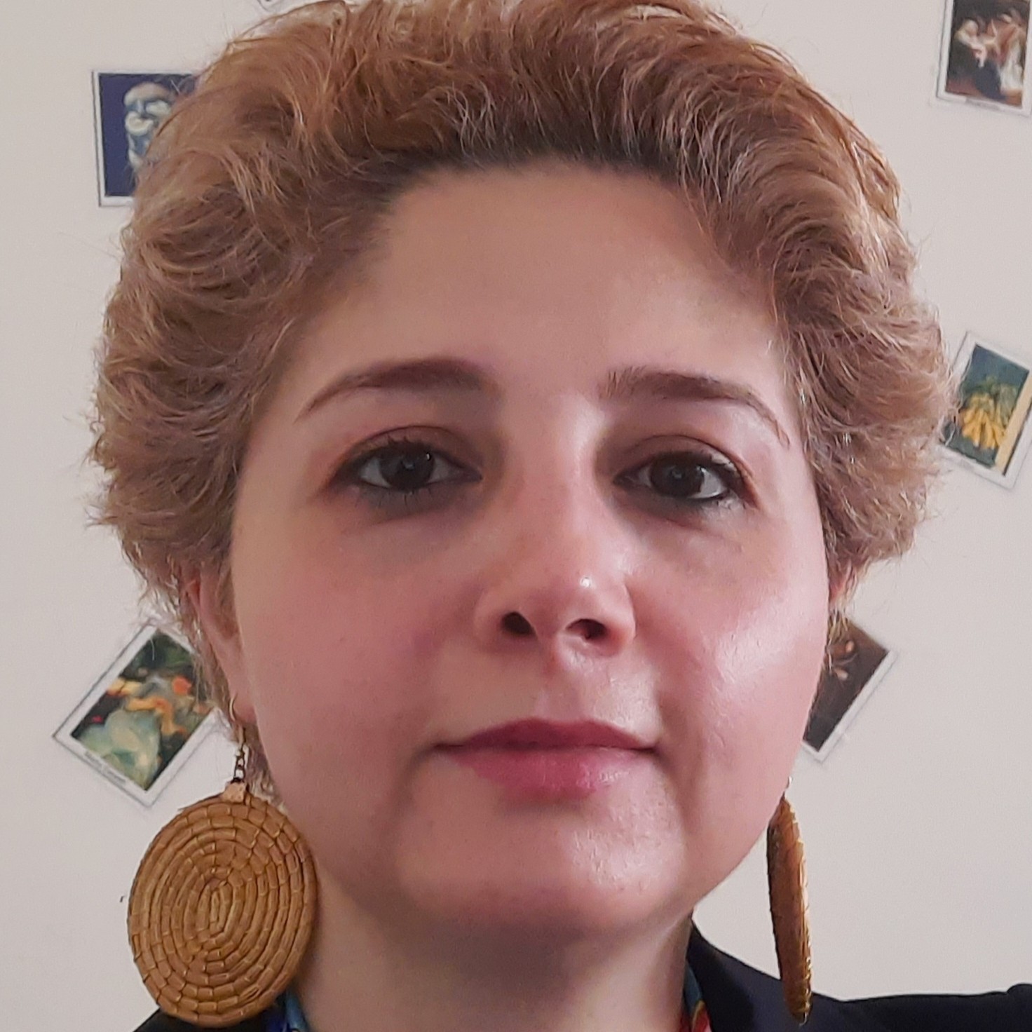 Encieh Erfani. Woman with curly blonde hair and large woven earrings looking at the camera. Small framed pictures adorn the wall behind her.